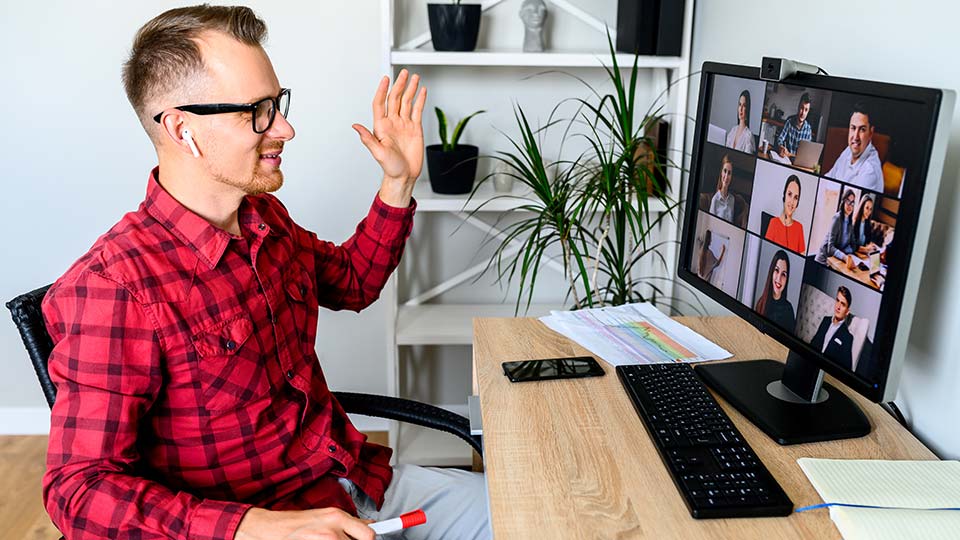 A young cheerful man greets with a gesture his coworkers in online video meeting, he sits at the office desk, a shelf with plants is on the background