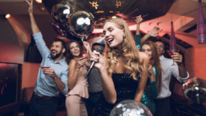 Young people have fun in a nightclub and sing in karaoke. In the foreground there is a woman in a black dress.