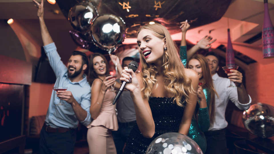 Young people have fun in a nightclub and sing in karaoke. In the foreground there is a woman in a black dress.